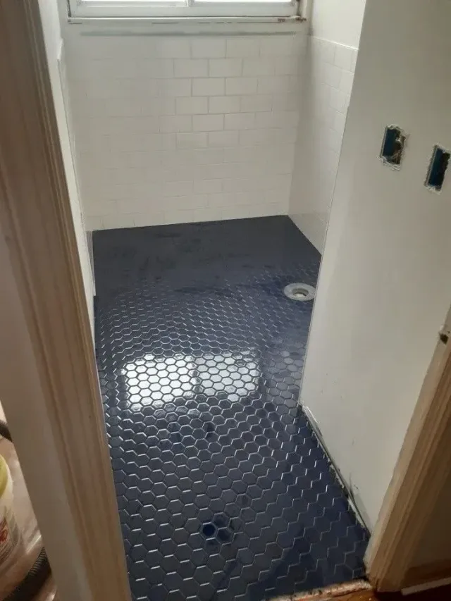 Small unfinished bathroom with dark hex tile floor, white tiled walls, and a window at the end.