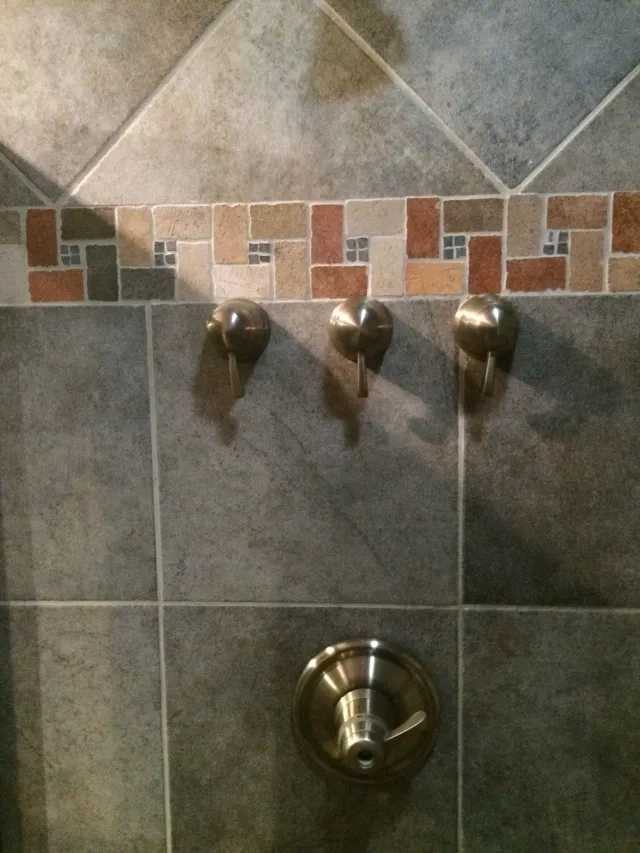 Shower wall with three metal hooks and a chrome knob on gray tiled background.