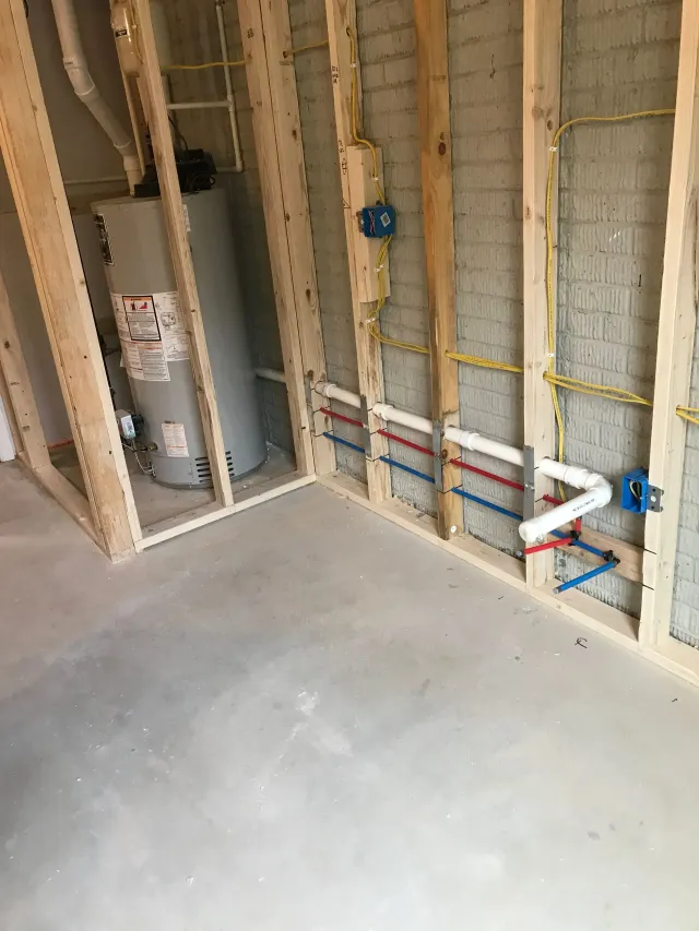 Unfinished basement wall with exposed framing, plumbing pipes, and a water heater beside a concrete floor